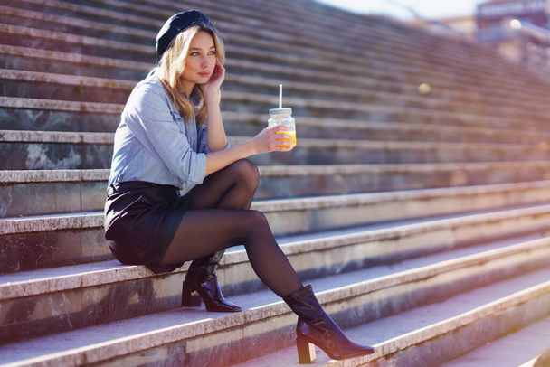 Ξανθιά γυναίκα με μπερέ, πίνοντας ένα φυσικό χυμό πορτοκαλιού σε ένα κρυστάλλινο ποτήρι, κάθεται σε κάποια βήματα. - Φωτογραφία, εικόνα