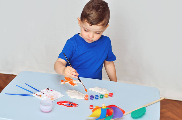 Un bambino piccolo ragazzo in una t-shirt blu dipinge figure in gesso magneti con vernici acriliche. - Foto, immagini