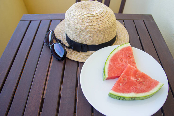 тарелка с ломтиками арбуза, шляпа и солнцезащитные очки на столе на террасе. Летняя концепция. Селективный фокус. - Фото, изображение