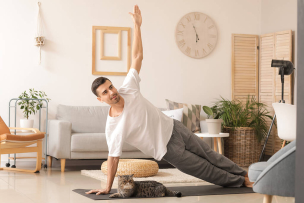 Junger Mann mit süßer Katze praktiziert Yoga zu Hause - Foto, Bild
