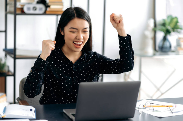 Happy positive successful asian young woman, stylishly dressed, office worker, freelancer or student sits at work desk, gesturing with her hands rejoicing in victory, success, good deal, big profit - Photo, Image