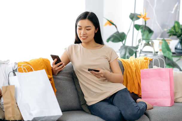 Positive stylish pretty young asian woman sits on sofa at home, uses smartphone and banking card for online payments, many paper bags with clothes near, enters card number, make online order, smiles - Photo, Image