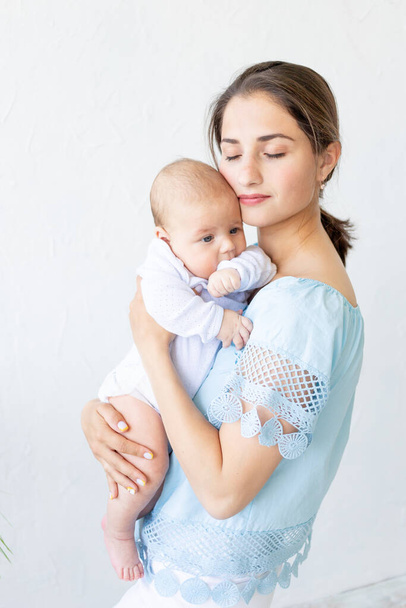 een jonge moeder met een pasgeboren baby houdt het zachtjes in haar armen, knuffelt en bewondert het thuis, het concept van een gelukkig gezin en de geboorte van kinderen - Foto, afbeelding