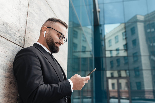 Happy mature businessman listening to music with wireless earphones and smartphone, leaning on business center wall - Photo, Image