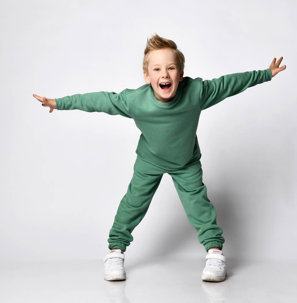 portrait of a cheerful little boy dressed in a green tracksuit. Cute baby posing on a white background - shouts with joy and spread his arms like wings. The concept of active childhood - Photo, Image