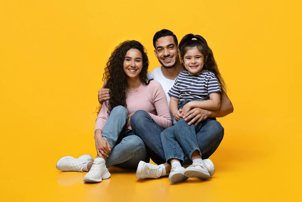 Happy middle eastern family sitting on floor and embracing over yellow background - Foto, Imagen