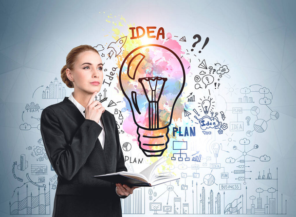 European attractive young woman in formal suit holding planner and pondering near light blue wall with colorful business idea lightbulb sketch. Concept of creativity, start up and brainstorming - Foto, imagen