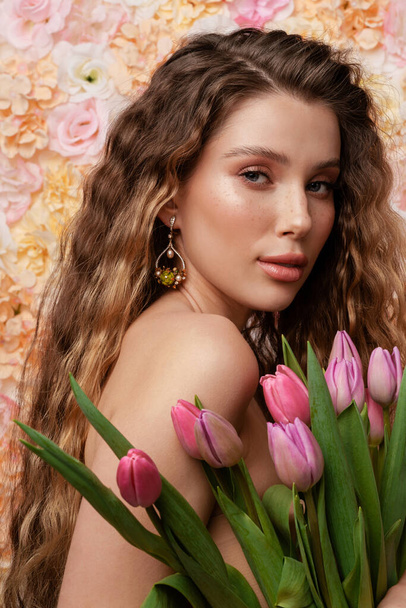 Half-naked young woman wearing earring posing with tulips isolated over flowers background - Фото, изображение