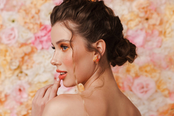 Half-naked young woman with earring posing and looking aside isolated over flowers background - Foto, Imagem