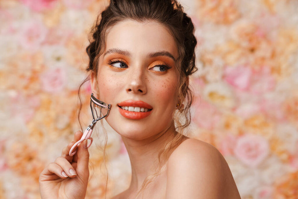 Half-naked pleased woman wearing earrings posing with eyelash curler isolated over flowers background - Foto, Bild