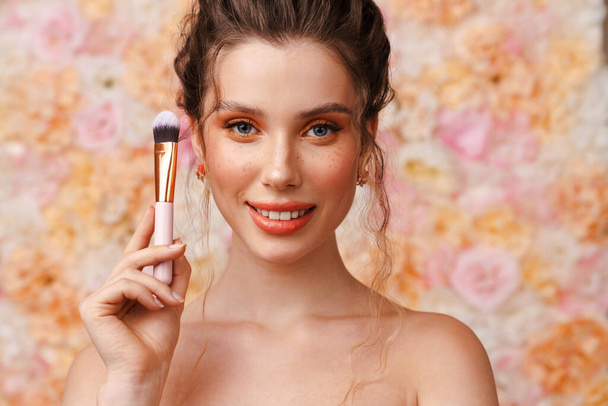 Half-naked young woman smiling while posing with powder brush isolated over flowers background - Photo, image
