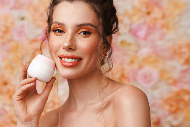 Half-naked young woman smiling while posing with face cream isolated over flowers background - Photo, Image