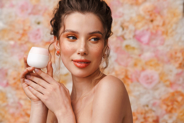 Half-naked young woman looking aside while posing with face cream isolated over flowers background - Photo, Image
