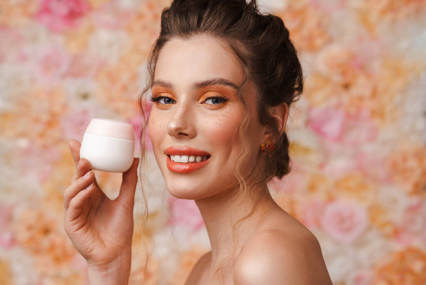 Half-naked young woman smiling while posing with face cream isolated over flowers background - Fotó, kép
