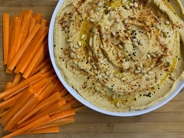 Hummus fatto in casa cosparso di olio d'oliva, decorato con pinoli, con bastoncini di carota su un piatto bianco. - Foto, immagini