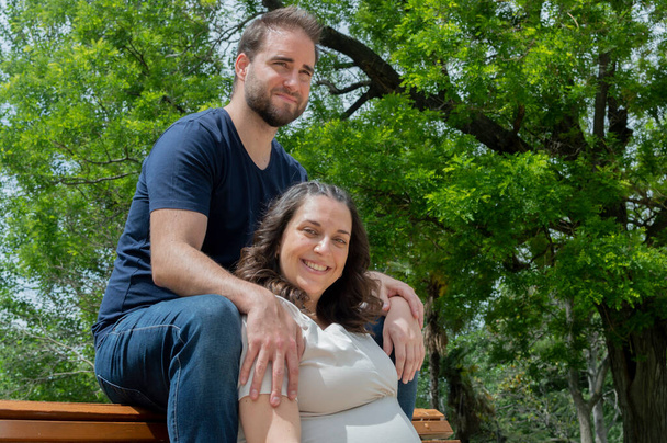 Zwanger paar zitten op een bank en poseren in een park - Foto, afbeelding