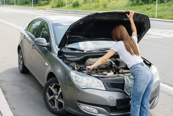 A young girl stands near a broken-down car in the middle of the highway and looks under the hood. Failure and breakdown of the car. Waiting for help. - Foto, immagini