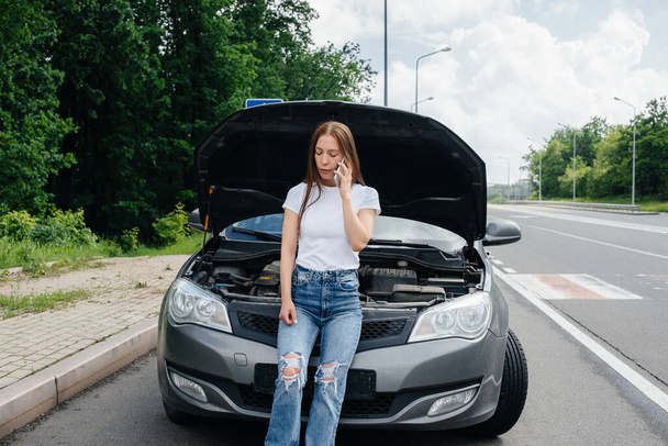 A young girl stands near a broken-down car in the middle of the highway and calls for help on the phone. Failure and breakdown of the car. Waiting for help. - Foto, Imagem