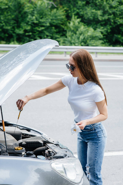A young girl stands near a broken-down car in the middle of the highway and checks the oil level in the engine. Failure and breakdown of the car. Waiting for help. - Foto, Bild