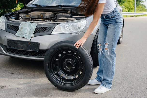 A young girl stands near a broken car in the middle of the highway and tries to change a broken wheel on a hot sunny day. Failure and breakdown of the car. Waiting for help. - Foto, immagini