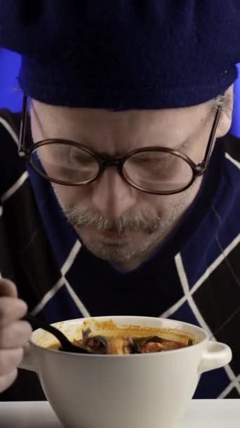Close-up van een man met een snor die Tom Yam eet en goedkeurend knipoogt. Verticale video. Verticaal. Blauwe achtergrond. - Video