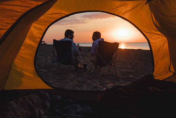 junges erwachsenes Paar, das dem Sonnenaufgang beim Kaffeetrinken im Campingstuhl zusieht. Lebensstil. - Foto, Bild