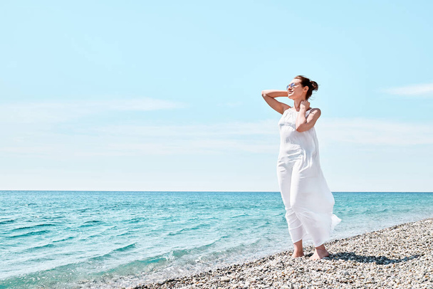 Happy woman in white dress walks in the pebble beach with crystal sea and blue sky and enjoys her vacation. Beach relax lifestyle. - Fotografie, Obrázek