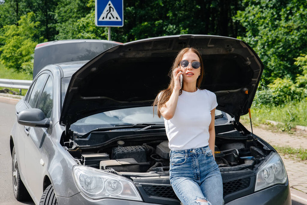 A young girl stands near a broken-down car in the middle of the highway and calls for help on the phone. Failure and breakdown of the car. Waiting for help. - Foto, immagini