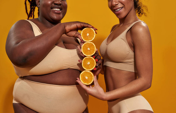 Cropped shot of two african american women in underwear smiling while holding halves of ripe juicy orange, standing isolated over orange background - Foto, Bild