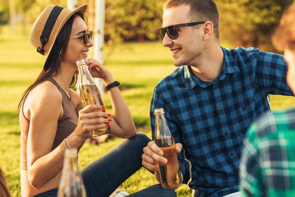 Beautiful young woman in a summer hat and sunglasses, communicates with a young man, drinking drinks on a picnic in the park on a sunny day - Foto, Imagem