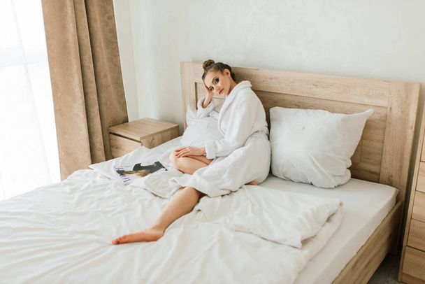 Woman in white robe stay near the window and on a bed in hotel room - Фото, изображение