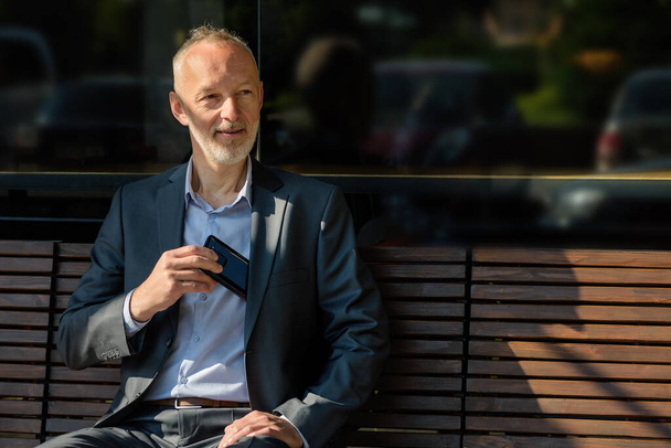 El hombre en un traje gris con un teléfono inteligente sentado en un banco. - Foto, imagen