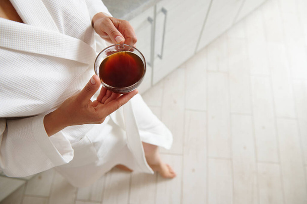 Vue du dessus de la tasse avec café dans les mains d'une femme en peignoir gaufré blanc debout à la cuisine de la maison, appuyé sur un comptoir de cuisine - Photo, image