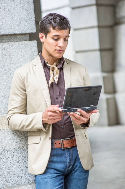 Vestirse con una chaqueta de color beige, una camisa de color café, vaqueros azules, una bufanda atada en el cuello, un joven guapo está mirando un pequeño ordenador y estudiando fuera. - Foto, Imagen