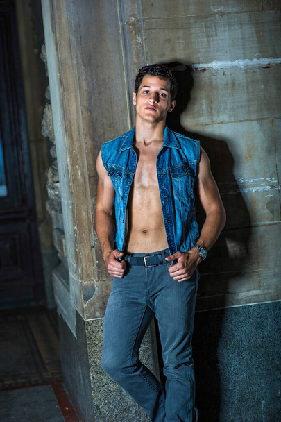 Dressing in a blue sleeveless Denim jacket, unbuttoned, a black pants , and standing in the front of a wall corner, a sexy young worker is taking a break outside, - Photo, Image
