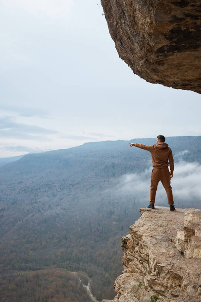 un uomo si erge su una roccia e punta il dito verso la valle, le montagne, la vista della foresta dalla scogliera - Foto, immagini