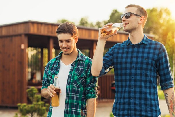 Two young male friends walking in the park in summer on a sunny day, talking and drinking beer drinks in nature - Foto, Imagem
