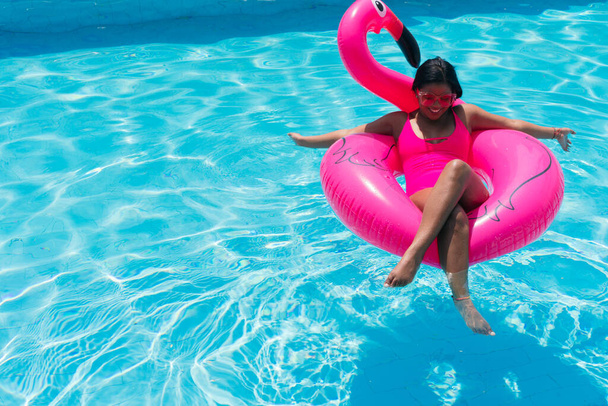 Smiling young woman in bikini relaxes on inflatable pink flamingo in swimming pool. Attractive woman in swimsuit lies in the sun on tropical vacation. Woman sunbathing at the resort. - Foto, Imagem
