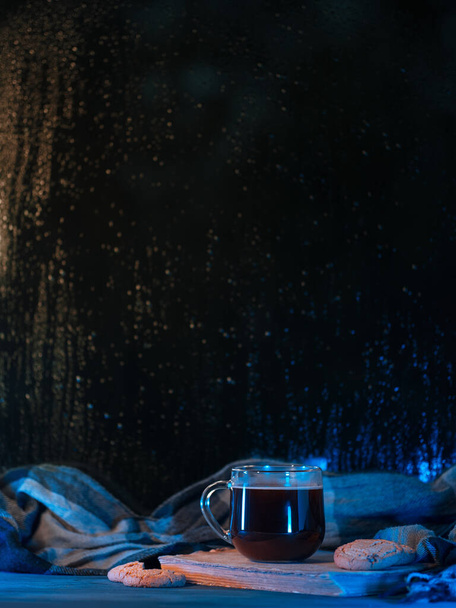 Ένα ποτήρι καφέ στο περβάζι του παραθύρου μια βροχερή νύχτα. Υγρό παράθυρο, παλιό βιβλίο, μπισκότα βρώμης - Φωτογραφία, εικόνα