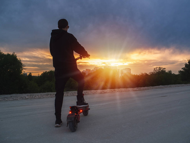 Un joven en un scooter eléctrico en el fondo de la puesta de sol. Transporte urbano moderno. - Foto, Imagen