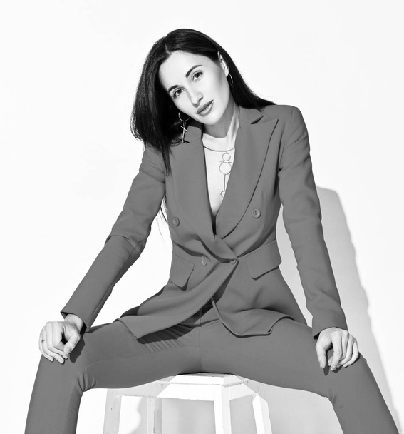 Black and white portrait of young brunette woman in official pantsuit sitting on a stool with her legs spread wide - Fotografie, Obrázek