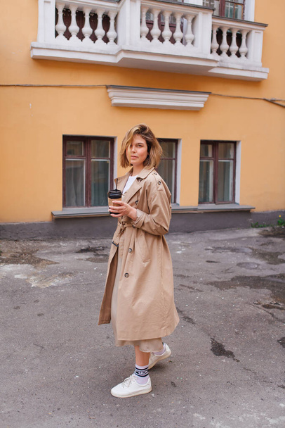 Junge lächelnde Millennial-Frau mit wilden Haaren im Herbstmantel spaziert mit einer Tasse Kaffee in die Nähe des alten Gebäudes. - Foto, Bild