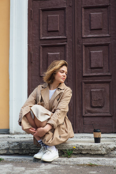 Young smiling millennial woman with wild hair dressed in an autumn coat sitting near the door of an old building with a cup of coffee to go. - Φωτογραφία, εικόνα
