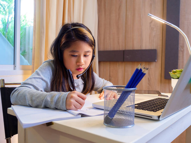  Asian girl in headphones sit at desk study online on laptop. Kid wear headset handwriting in notebook learning using internet lessons on quarantine. Girl student learning virtual internet online class from school due to covid pandemic. - Photo, Image