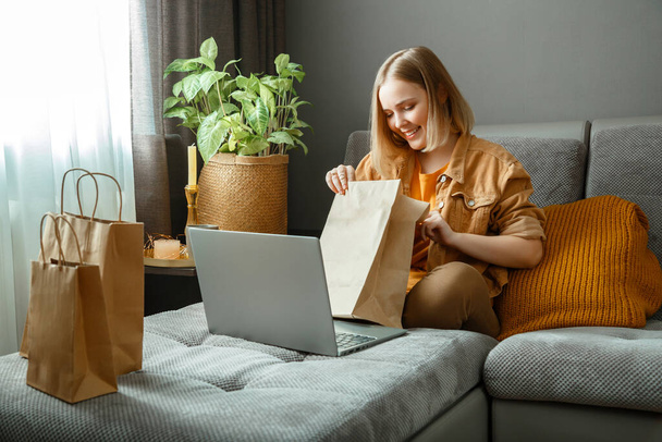 Happy young woman do unpacking online orders goods or food. Online Shopping, ordering delivery. Teenager girl relax on sofa considering purchases with laptop. Mock up paper bags - Fotografie, Obrázek