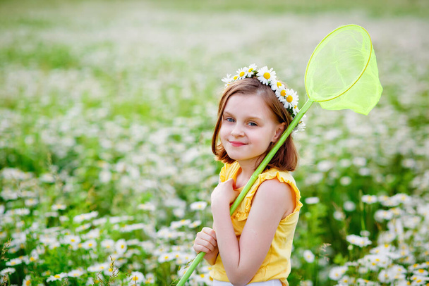 Porträt eines süßen Mädchens in einem gelben T-Shirt mit einem geblümten Gänseblümchenkranz auf dem Kopf. Ein Kind steht mit einem Schmetterlingsnetz in einem Kamillenfeld. - Foto, Bild