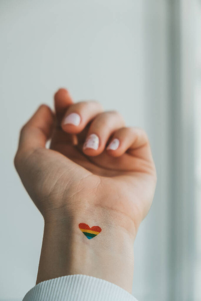 Primer plano de la joven mujer hippie milenaria caucásica con una bandera de arco iris en forma de corazón pintado en la muñeca. Tatuaje de color arco iris, símbolo del mes del orgullo LGBTQ. Igualdad de derechos - Foto, imagen