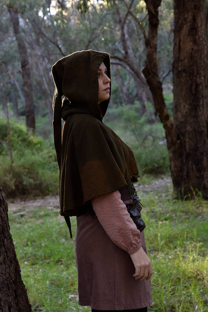  πορτραίτο της κοκκινομάλλας που φορούσε φανταστικά μεσαιωνικά ρούχα ενός περιπλανώμενου τυχοδιώκτη. φυσικό φως σε δασικό τοπίο. - Φωτογραφία, εικόνα