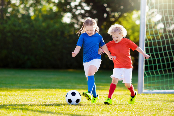 Kids play football on outdoor field. Children score a goal at soccer game. Girl and boy kicking ball. Running child in team jersey and cleats. School football club. Sports training for young player. - Фото, изображение