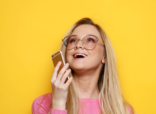 Happy pretty lady with blond hair and blue eyes using mobile phone, standing against yellow background. - Fotó, kép